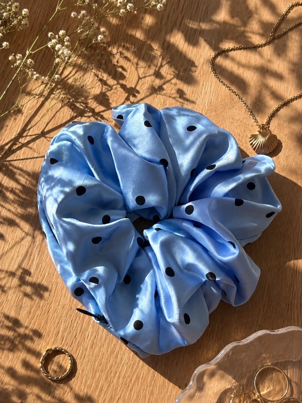 Light blue Carimé scrunchie with black dots on a wooden table in sunlight, next to a gold necklace