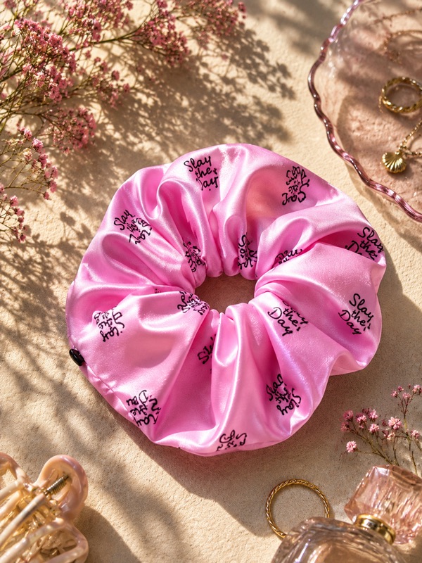 Pink Carimé scrunchie with black “Slay the Day” lettering on a sand-coloured surface, surrounded by dried flowers