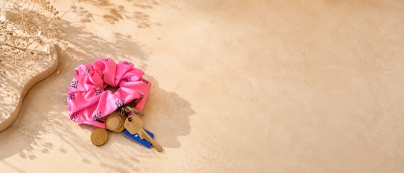 Wide shot of the pink scrunchie with keys and coins on pale sand, with open space to the right