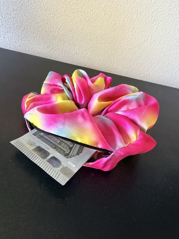 Das Gradient-Scrunchie mit geöffnetem Reißverschluss, ein gefalteter Geldschein ragt heraus